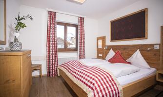 A cozy bedroom with a double bed and wooden furniture. Red checkered fabrics and a large window front create an inviting atmosphere.