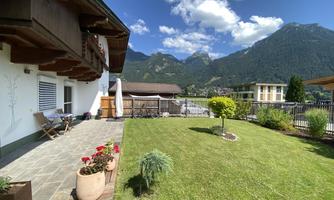 A sunny terrace with a well-kept garden and a view of the mountains. Blue skies and white clouds complete the idyllic atmosphere.
