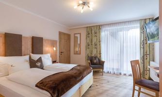 A modern hotel room with two beds, a cozy armchair, and a desk. Large windows ensure plenty of natural light.
