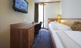 A modern hotel room with a double bed and a desk. A television hangs on the wall, and the room has large windows with daylight.
