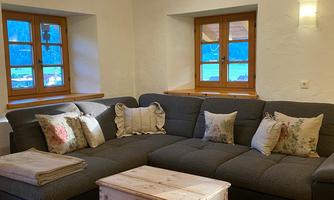 A cozy living room with a gray sofa and cushions. A wooden table stands in the middle and there are large windows looking outside.