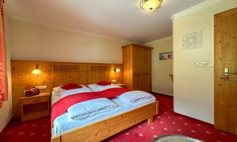 A cozy bedroom with wooden furniture and a red carpet. The room is bright and inviting with a large double bed and a continuous wall lamp.