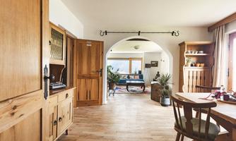 A bright and inviting living room with wooden furniture and a large window. The space is decorated with plants and a rug, creating a cozy atmosphere.