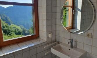 A modern bathroom with a sink and a large mirror. Through the window, there is a view of the green mountain landscape.
