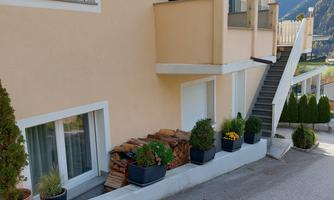A bright residential building with a balcony and two floors. In front of the house, there are several plant pots with flowers and small trees.