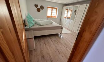 A bright bedroom with a large bed and a white wardrobe. The walls are plain, and there are large windows that let light in.