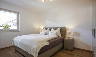 A modern bedroom with a large bed and elegant bedding. The room has light walls, a window, and minimalist design.