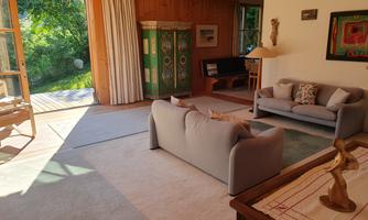 A bright and inviting living space with a cozy sofa and a wooden table. Large windows provide a view of the green surroundings.