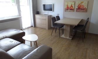 A bright living room with a cozy sofa, a table, and dining chairs. Pictures of fruits hang on the wall, and a television is also available.