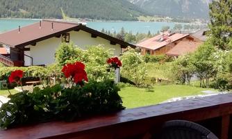 A beautiful view from a balcony of a lake, surrounded by mountains and green landscape. In the foreground, blooming flowers and a table with chairs can be seen.