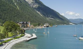 A picturesque lake surrounded by mountains and trees. Sailboats can be seen at the shore, and a beautiful path invites you to take a stroll.