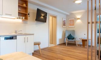 A modern living environment with a functional kitchen and hardwood floors. The space is bright and inviting, featuring a cozy armchair and appealing wall decorations.