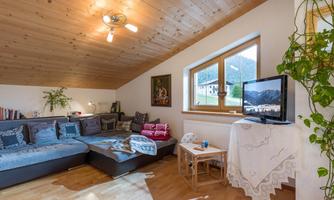 A cozy living room with a gray sofa, a low table, and a television. Large windows provide plenty of daylight and a view of the mountains.