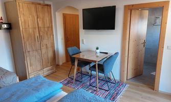 A cozy room with two beds, a table, and chairs. The room is bright and modernly furnished, with wooden furniture and a television on the wall.