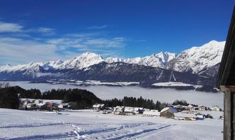A picturesque winter landscape with snow-covered mountains and a clear blue sky. In the foreground, snow-covered houses and a icy path can be seen.