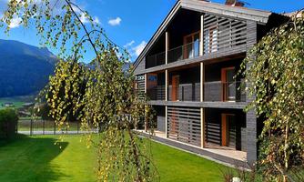 A modern wooden house surrounded by green grass and trees. In the background, mountains and a blue sky are visible.