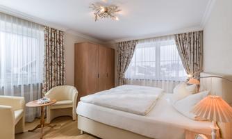 A cozy bedroom with a large bed and two chairs. The windows are adorned with curtains, and the room is bright and invitingly designed.