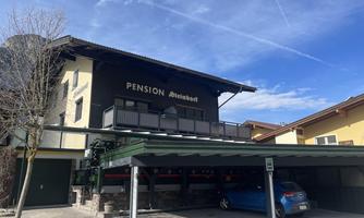 A guesthouse named "Pension Steinbock" is in the foreground. The building has a balcony and a car is visible in the covered parking garage.