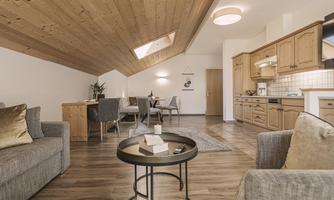 A cozy living space with a wooden beam ceiling and bright interior. The furnishings include a kitchen, a dining table, and comfortable sofas.