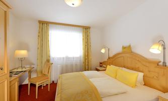 A bright room with wooden furniture and a large window. The bed is decorated with yellow pillows and there is a small desk.