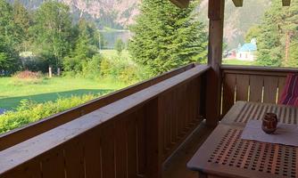 A balcony with a wooden railing and a view of a green landscape and mountains. The sun is shining, and it is a calm, natural environment.