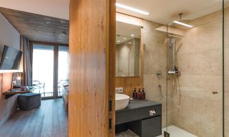 A modern bathroom with a glass shower and a large sink. In the background, there is a bright bedroom with wooden details and windows visible.