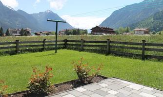 A beautiful garden with green grass and flowers. In the background, mountains and some buildings can be seen.