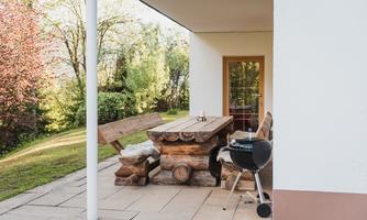 A cozy outdoor dining area with a large wooden table and a grill station. In the background, there are green trees and an inviting garden landscape.