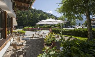 A cozy outdoor area with tables and chairs under a sun umbrella. In the background, green trees and a picturesque landscape can be seen.