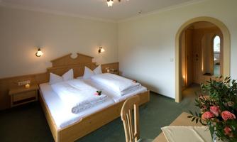 A cozy bedroom with a large double bed and white bed linen. In the room, there is a table with flowers and a wooden chair.