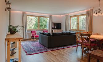 A cozy living room with a gray sofa and a wooden table. Large windows offer a view of the green surroundings.