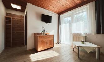 A bright room with a wooden floor and wooden ceiling. There is a window with curtains, a small table, and a wardrobe against the wall.