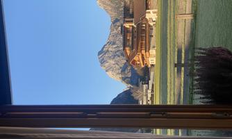 View from a window of a picturesque mountain landscape. In the foreground, there is a house and a meadow under a clear blue sky.