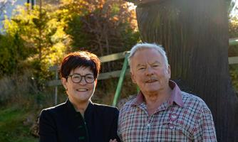 An older couple is standing outdoors, laughing in front of a tree. In the background, colorful autumn leaves can be seen.