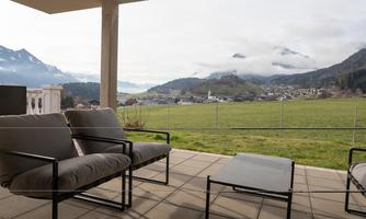 A cozy terrace with modern furniture that offers a view of spectacular mountains and a green landscape. The clouds are drifting across the sky, giving the scene a tranquil atmosphere.