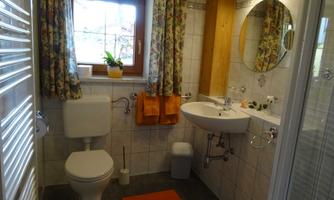 A small, bright bathroom with a window that shows a plant. There is a toilet, a sink, and a shower cabin, as well as tasteful curtains.