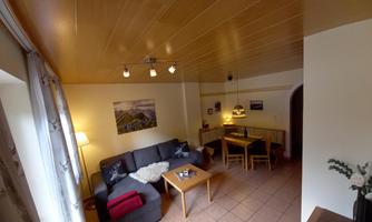 A cozy living room with a black sofa and a wooden table. In the background, a dining area and beautiful decoration can be seen.