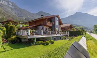 A modern house with a large terrace and garden, surrounded by mountains. The sky is clear and the landscape is green and well-maintained.