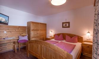 A cozy bedroom with a wooden double bed and red pillows. The walls are bright, and there is a desk corner with a wooden chair.