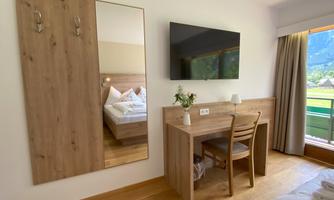 A modern room with a wooden desk, a chair, and a large mirror. A TV is mounted on the wall, and there is a balcony with a view of nature.