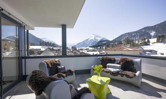 A modern balcony with comfortable chairs and a small table surface. The view shows snow-covered mountains and a clear sky.