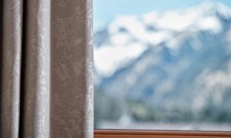 A curtain at the window with a view of blurred mountains in the background. The sky is clear and blue.
