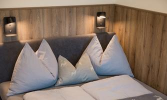 A cozy bed with simple, pointed pillows is placed in front of wooden paneling. Stylish wall sconces are mounted on the wall.