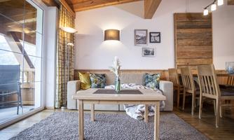 A cozy living room with a bright sofa, a wooden table, and a decorative plant. Large windows let in plenty of daylight and create an inviting atmosphere.