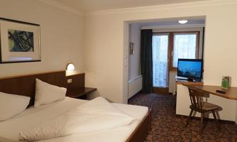 A simple hotel room with a double bed and a desk. In the background, you can see a television and large windows with a view outside.