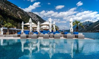 A luxurious pool with comfortable loungers and sunshades, surrounded by mountains. The sky is clear and the scenery is breathtaking.