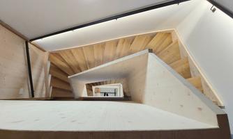 A modern wooden staircase with a spiral design. The staircase features light-colored steps and minimalist lighting.