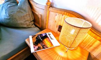 A cozy bedroom with a wooden bed, a pillow in soft colors, and a bedside table. On the table, there is a magazine next to a modern lamp.