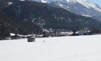 A vast, snow-covered landscape with mountains in the background. In the foreground are a few buildings and a person walking through the snow.