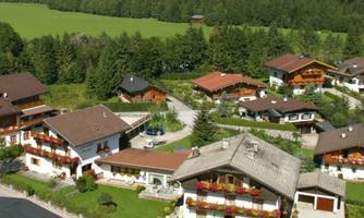 A picturesque residential settlement nestled amidst green fields and trees. The houses feature typical alpine architecture with balconies and blooming flower boxes.